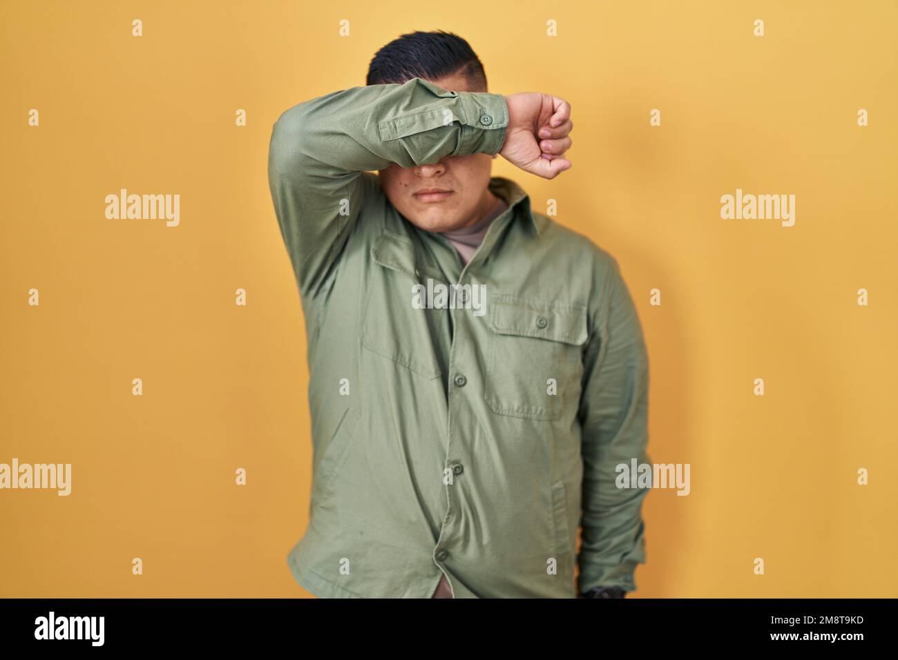 Hispanic young man standing over yellow background covering eyes with ...