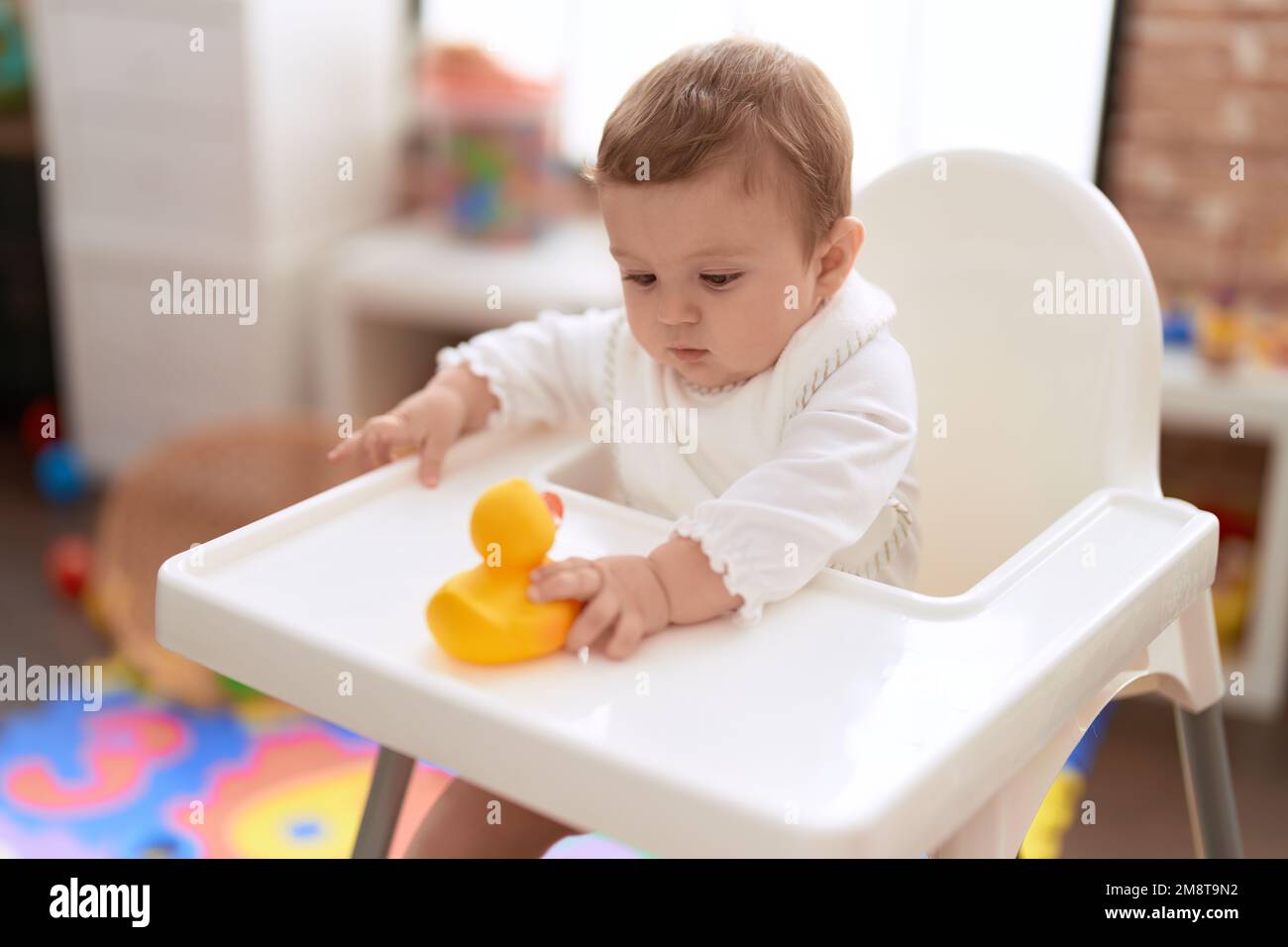 Adorable toddler sitting on baby highchair holding rubber duck toy at kindergarten Stock Photo