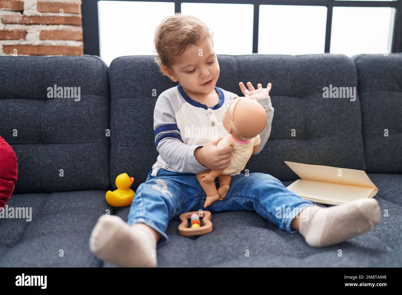 Adorable caucasian boy playing with baby doll sitting on sofa at home ...