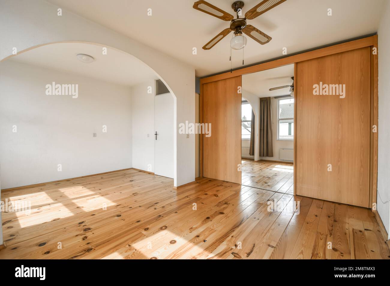 an empty room with wood flooring and ceiling fan in the middle part of the room is bright white walls Stock Photo