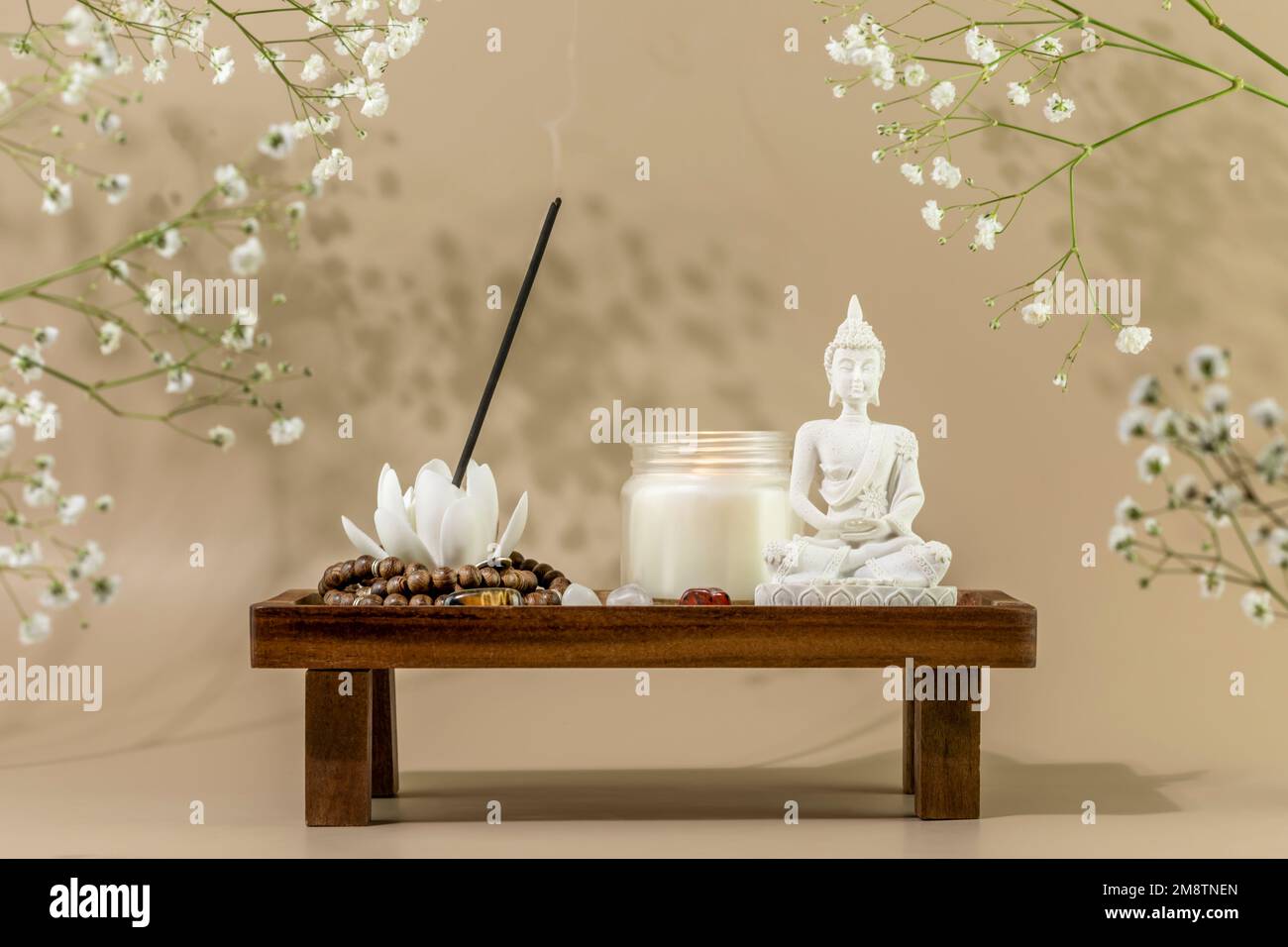 Self care still life with Buddha statue, candle, aroma stick on wooden podium with white flowers. Asian spa ritual procedure. Concept of Mental health Stock Photo