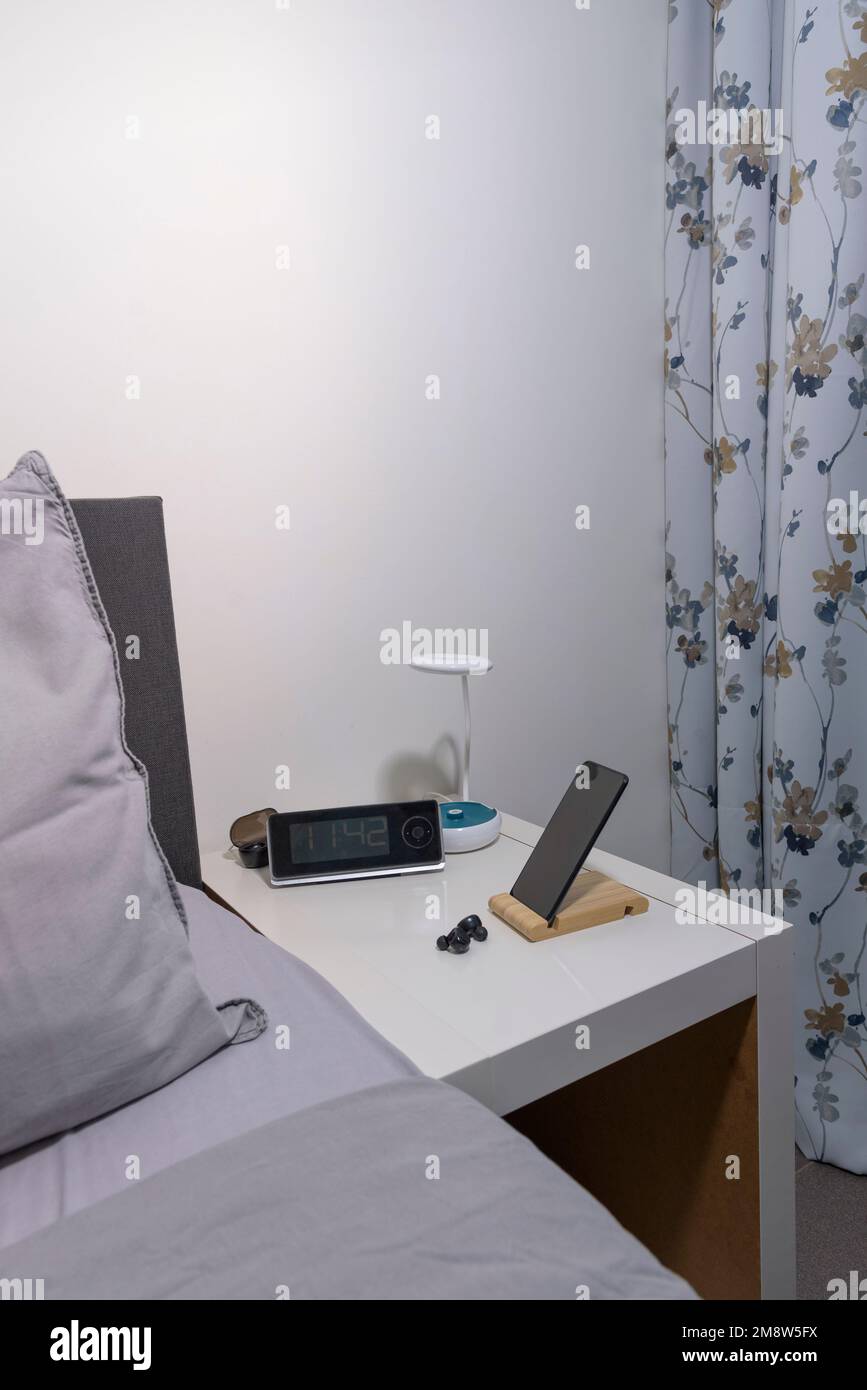 Smartphone on the bedside table with lamp, headset, clock and fragment of bed. Vertical view Stock Photo
