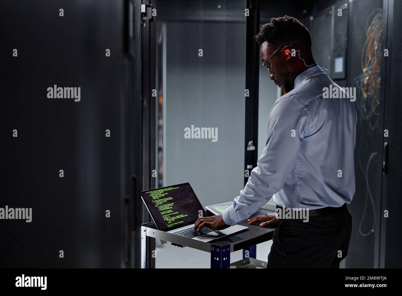 Side view portrait of black man as network engineer using laptop while setting up servers in data center Stock Photo