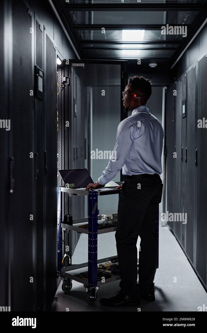 Vertical full length portrait of black man as network engineer using laptop and setting up servers in data center Stock Photo