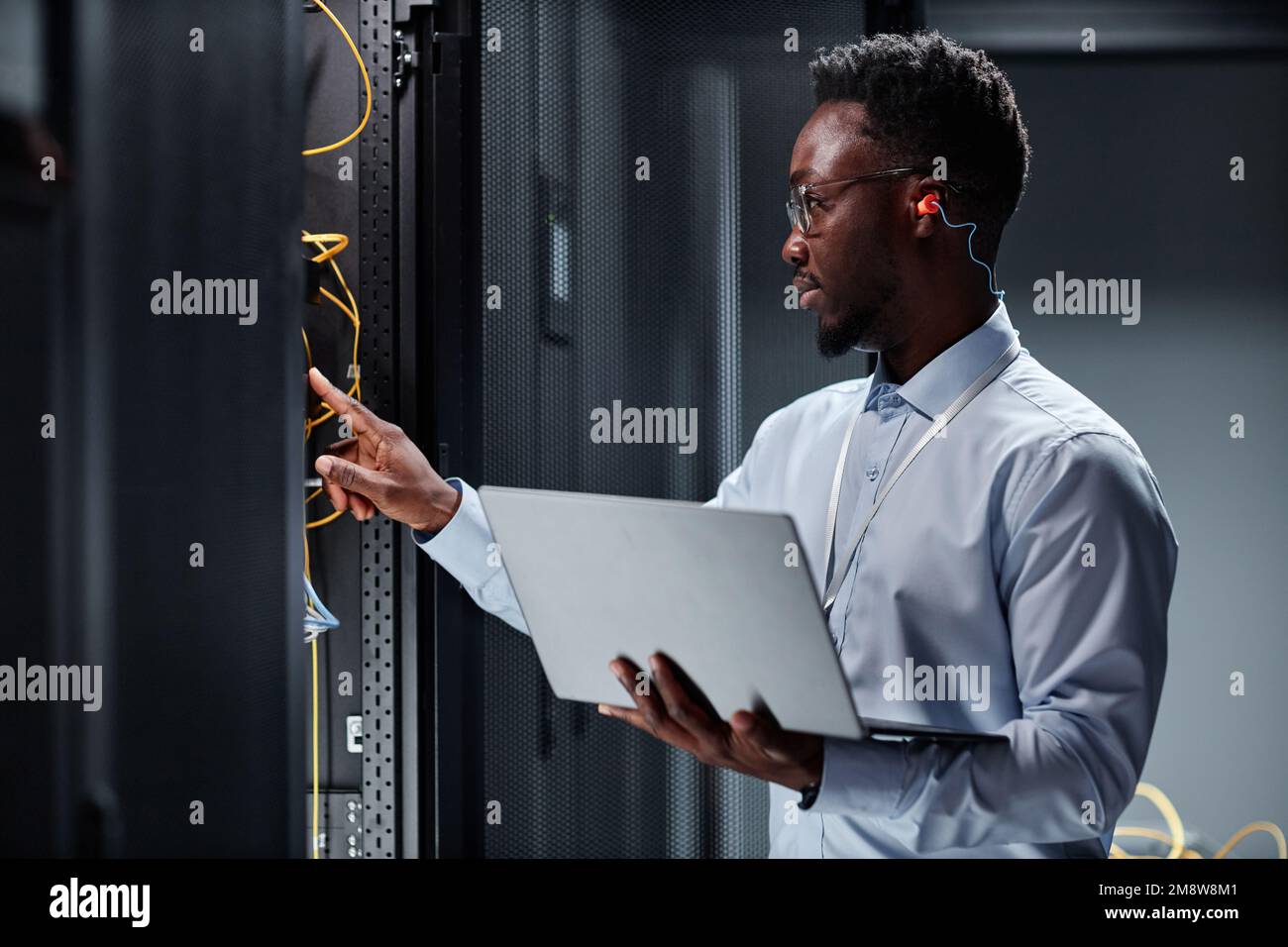 Side view portrait of young black man as network engineer working with servers in data center and holding laptop Stock Photo