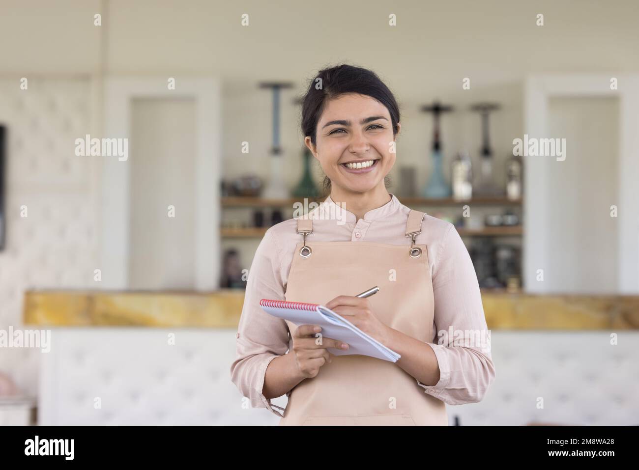 Happy beautiful Indian waitress in uniform apron holding pen Stock
