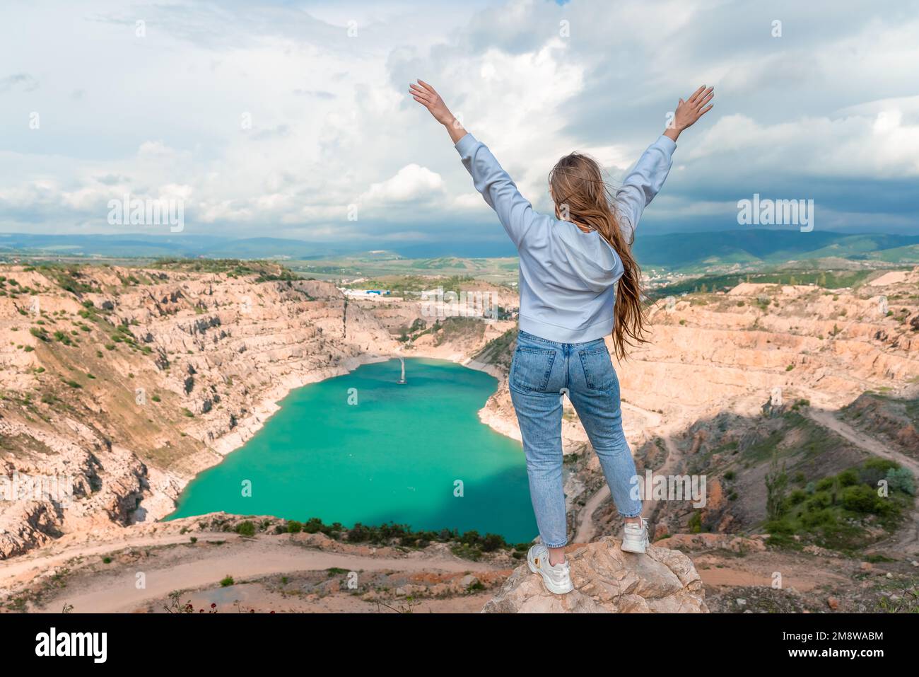 Girl on top of a mountain with raised hands, view of the azure lake in the form of a heart behind Stock Photo