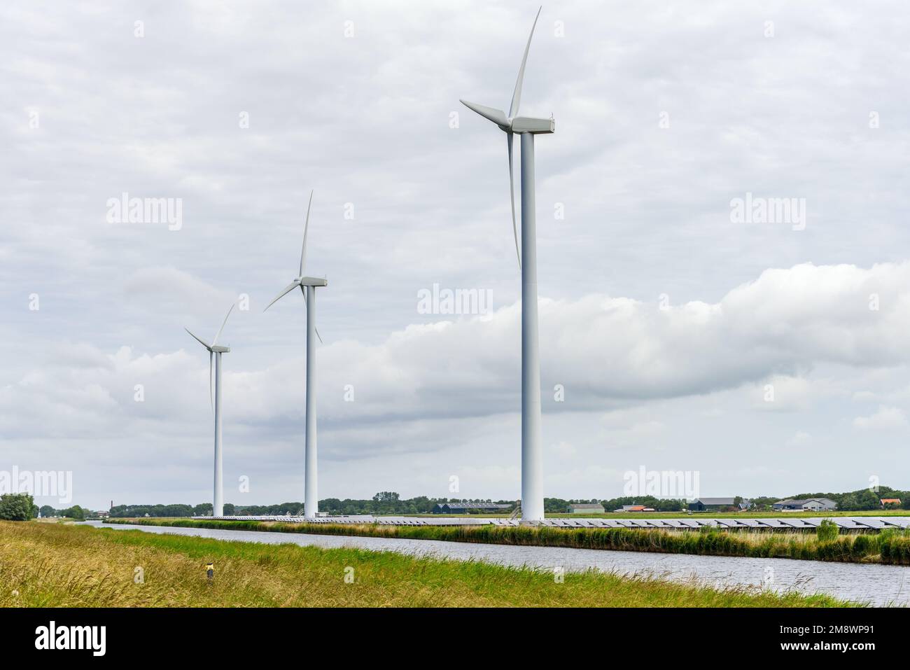 Wind turbines along a canal in the countryside of Netherlands on a cloudy summer day. Rows of solar panels are at the foot of the turbines. Stock Photo
