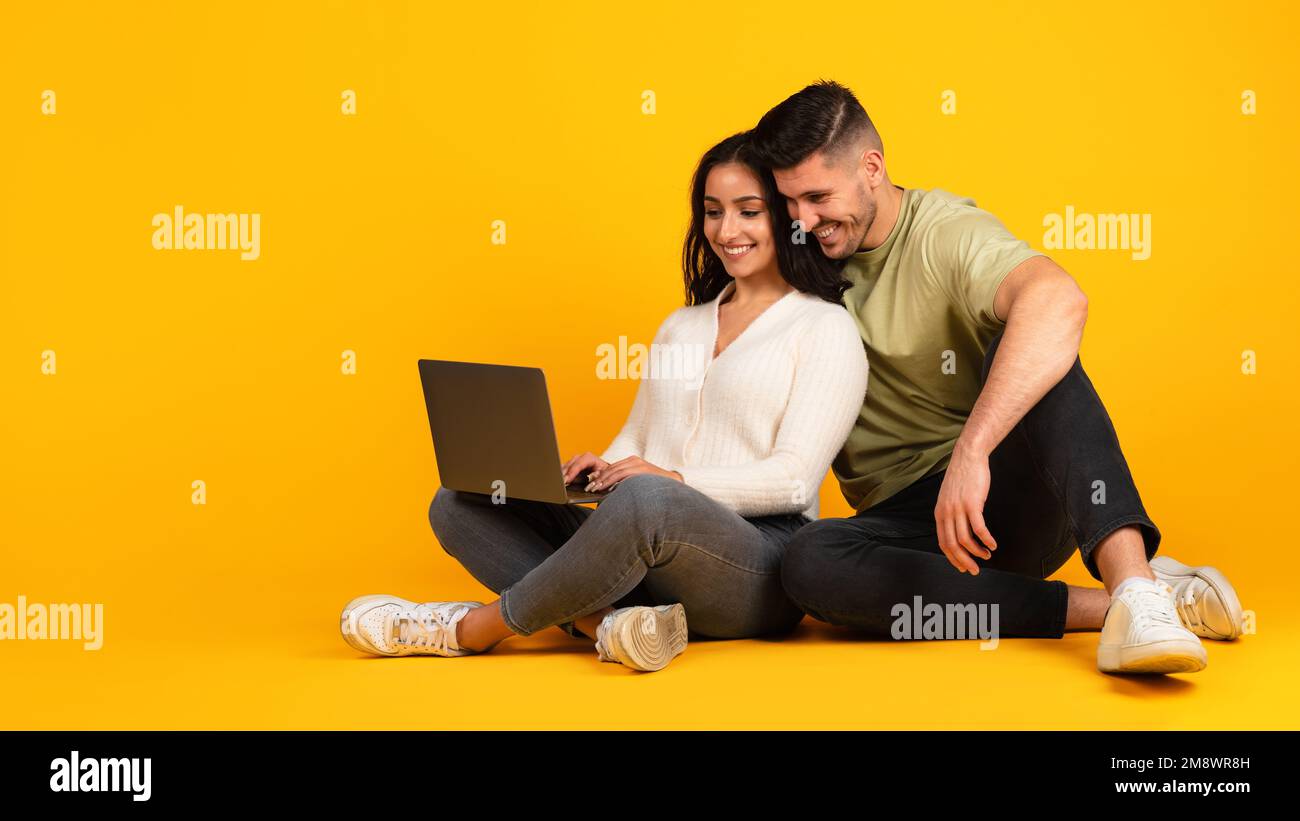 Smiling millennial middle eastern guy and woman in casual sit on floor typing in computer Stock Photo