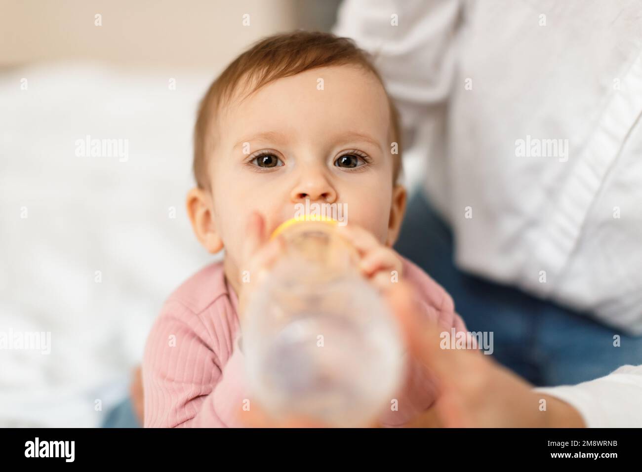 Cute infant baby girl drinking water from bottle, sitting on bed with