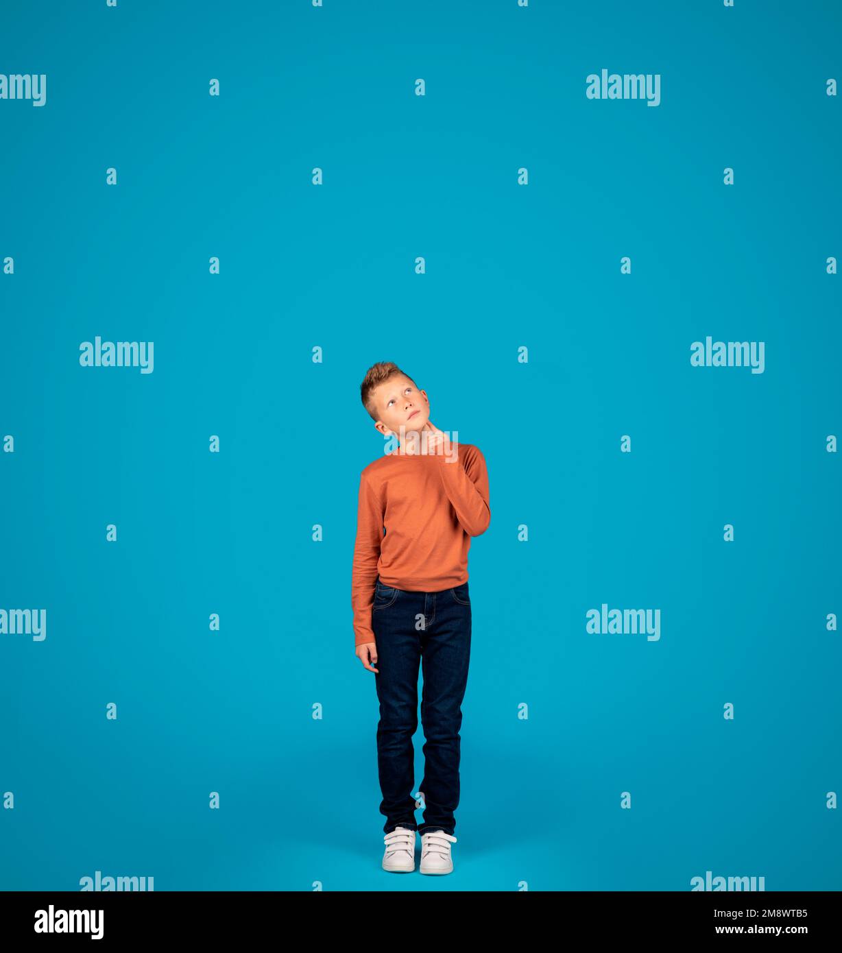 Thoughtful Preteen Boy Looking Up While Standing Over Blue Studio Background Stock Photo