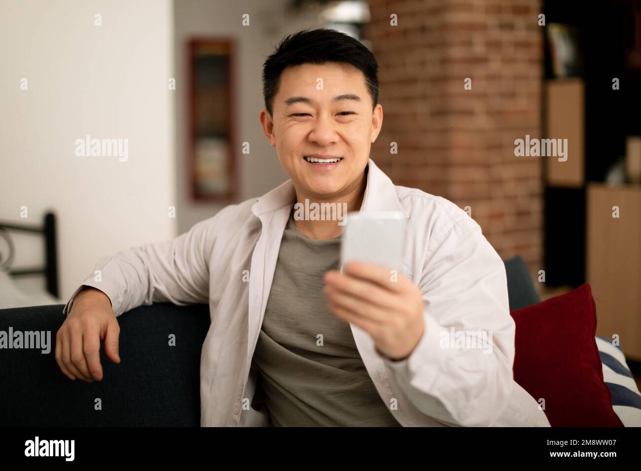 Happy mature asian man in casual watching video or chatting on smartphone, resting on sofa in living room interior Stock Photo
