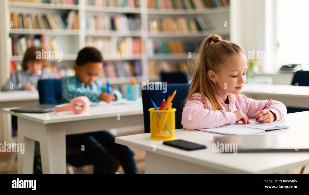 Diverse kids sitting at tables in classroom at primary school or kindergarten, writing or drawing in notebooks Stock Photo