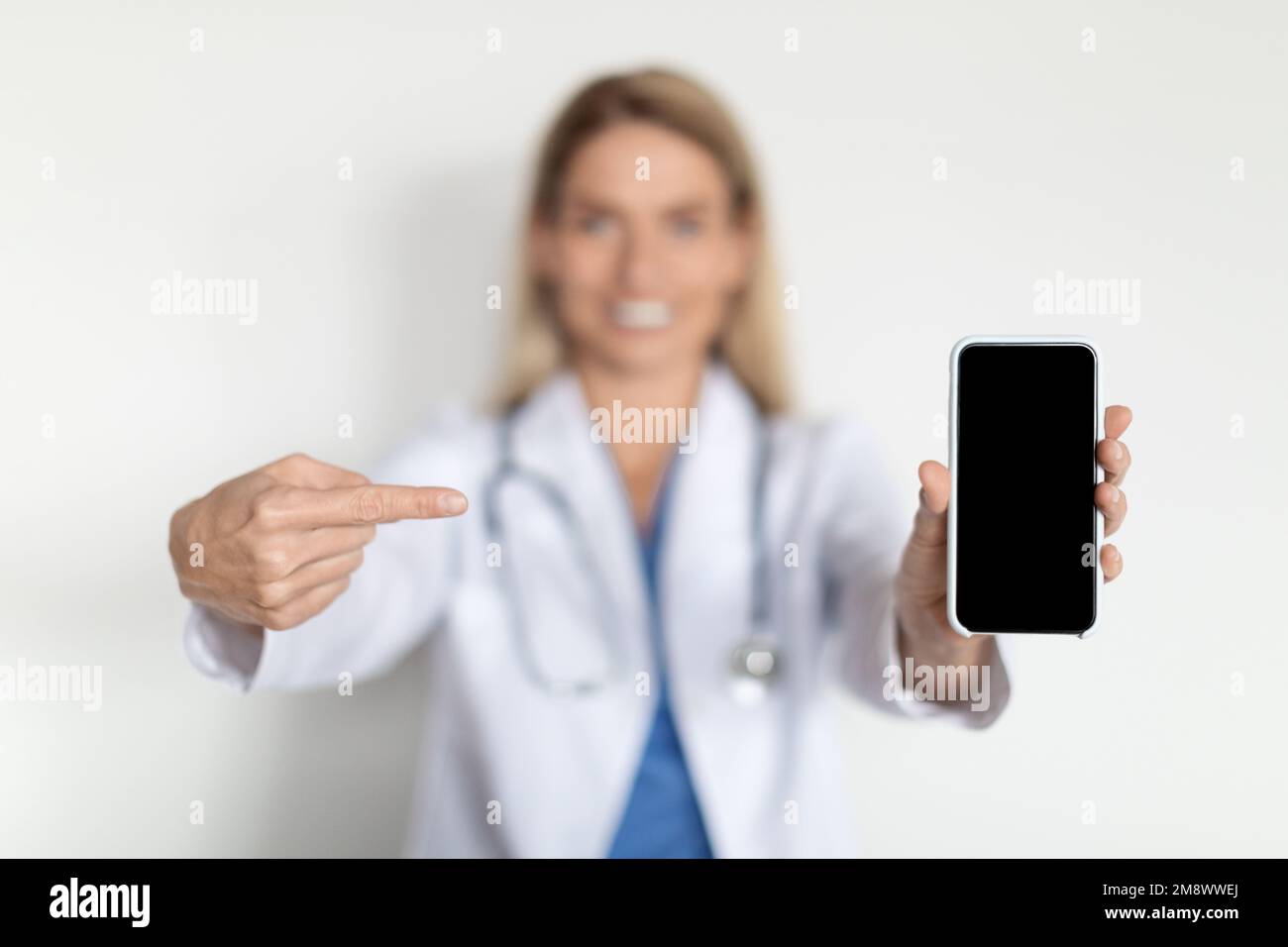 Medical Ad. Smiling Female Doctor Wearing Uniform Pointing At Blank Smartphone Stock Photo