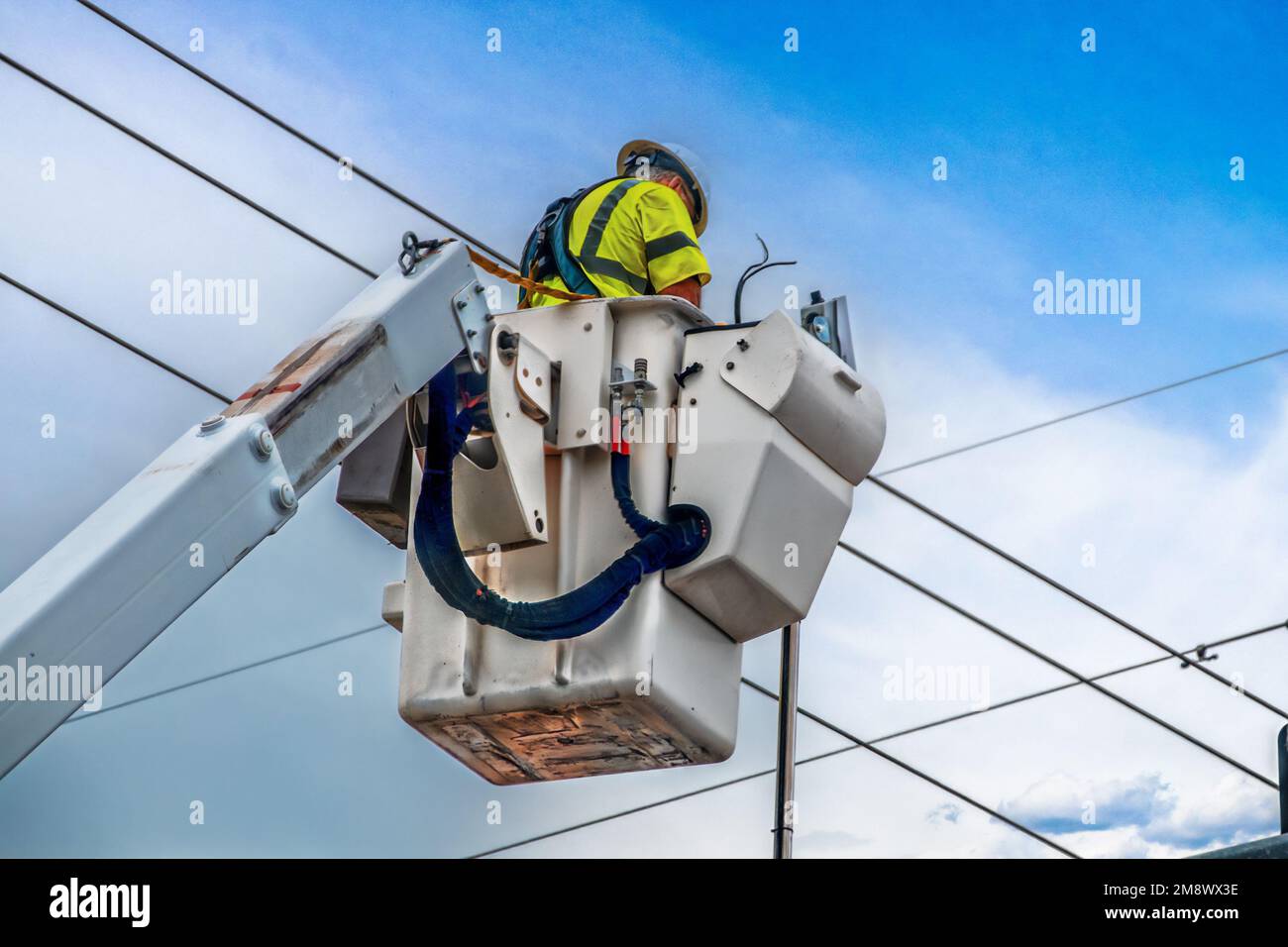 Electrician workman in green safety vest and helmet in crane lift working on highlines or traffic lights high in the air with sky background Stock Photo