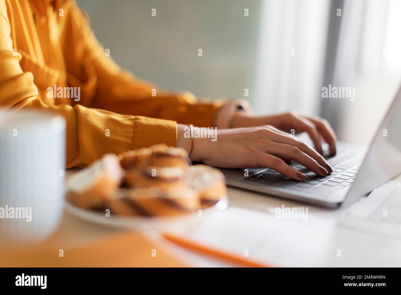 Female Hands Typing On Laptop Keyboard While Sitting At Table In Kitchen Stock Photo