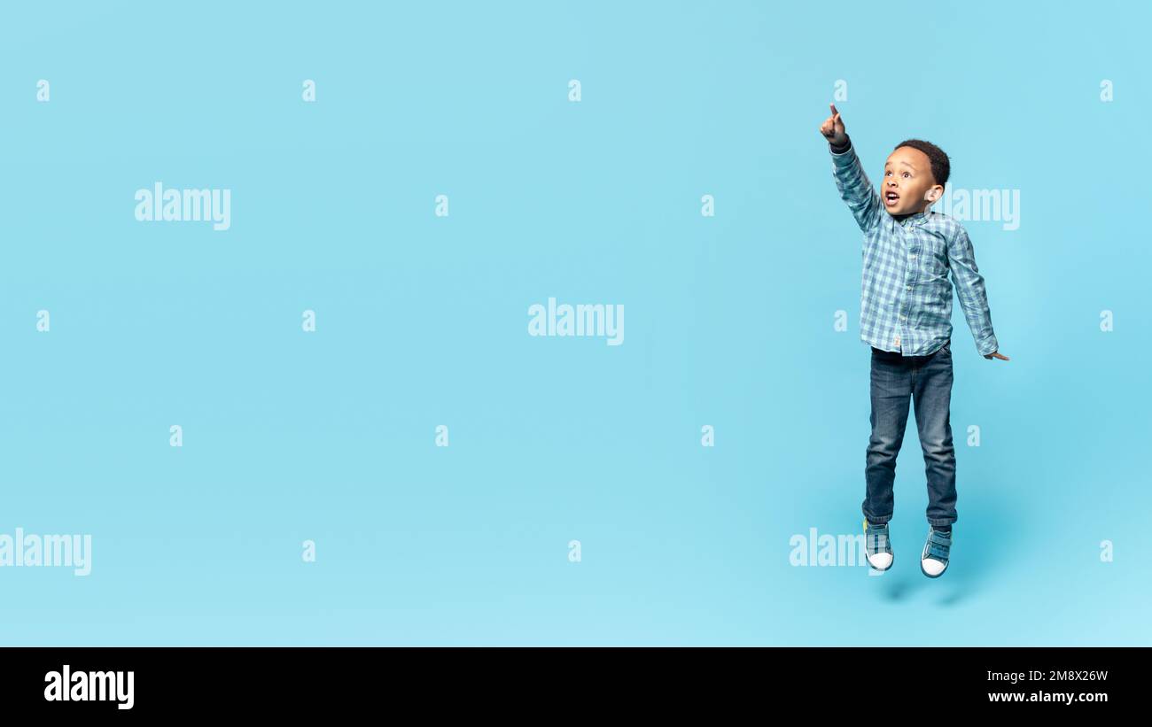 Emotional little african american boy jumping up and pointing at copy space for advertisement, blue background, panorama Stock Photo