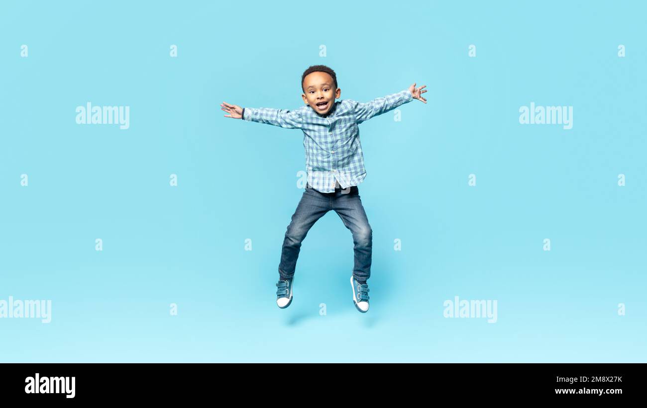 Happy little african american boy posing in mid air, jumping and spreading arms over blue studio background, full length Stock Photo