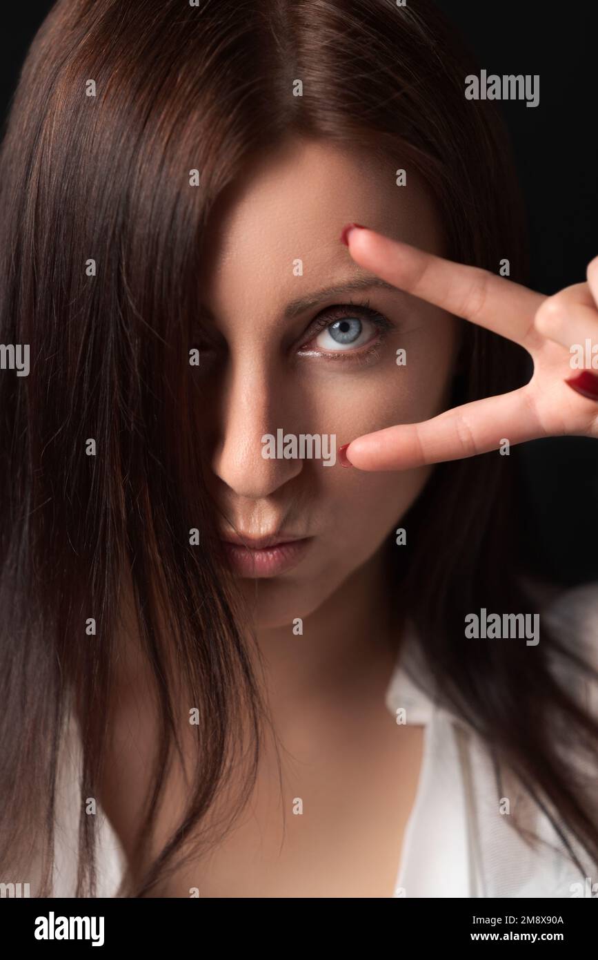 Portrait of serious adult woman holding two fingers near eye, showing victory or peace gesture and looking at camera. Sulking nice brunette Stock Photo