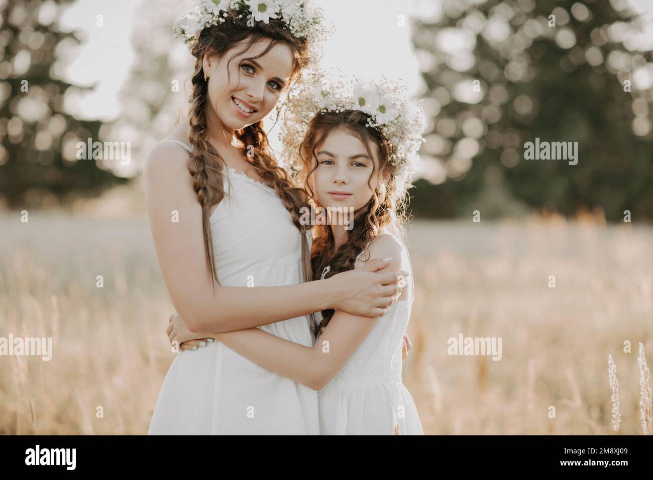 happy mom and daughter in white dresses with floral wreaths and boho style braids in summer in a field in nature. Added a small grain, imitation of fi Stock Photo