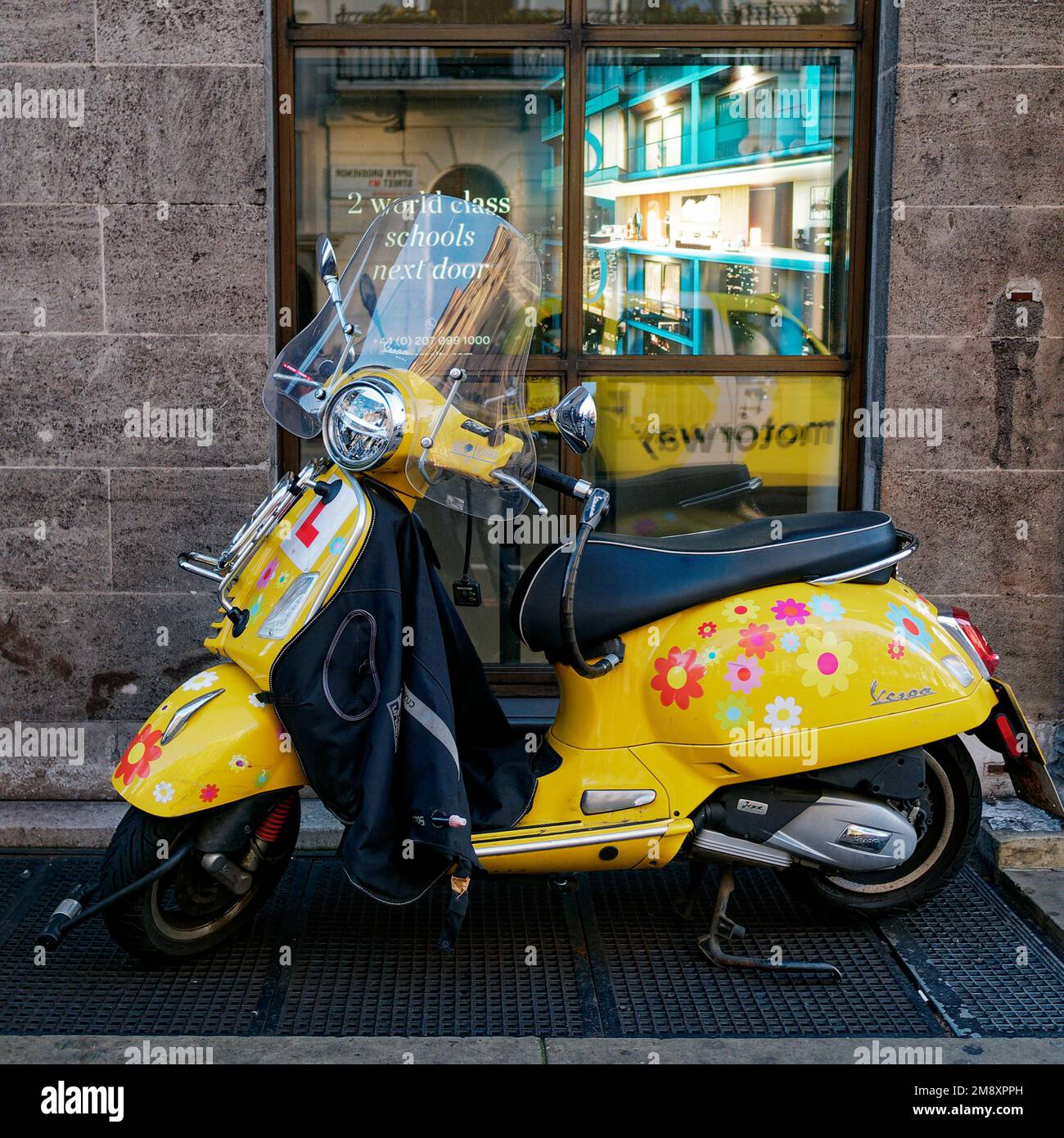 Yellow Vespa aka Scooter with flower design and learner plates in front of a window reflecting a yellow cab aka taxi. London, England Stock Photo