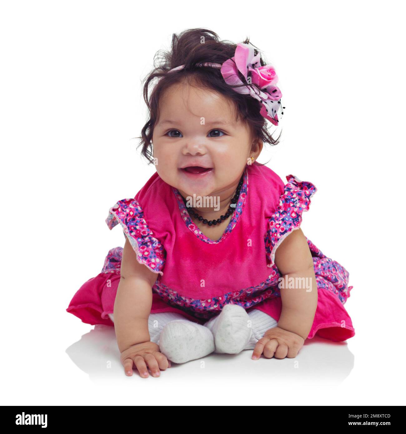 Innocent, cute and happy baby girl in a studio with a floral, beautiful and flower outfit and headband. Happiness, smile and infant child sitting and Stock Photo