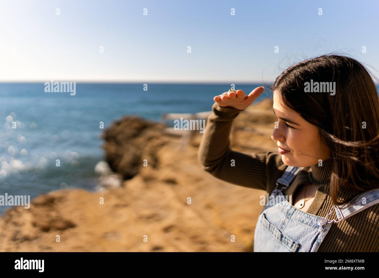 Selective focus side view of ethnic female protecting eyes from sun with hand on beach near calm sea in daylight Stock Photo