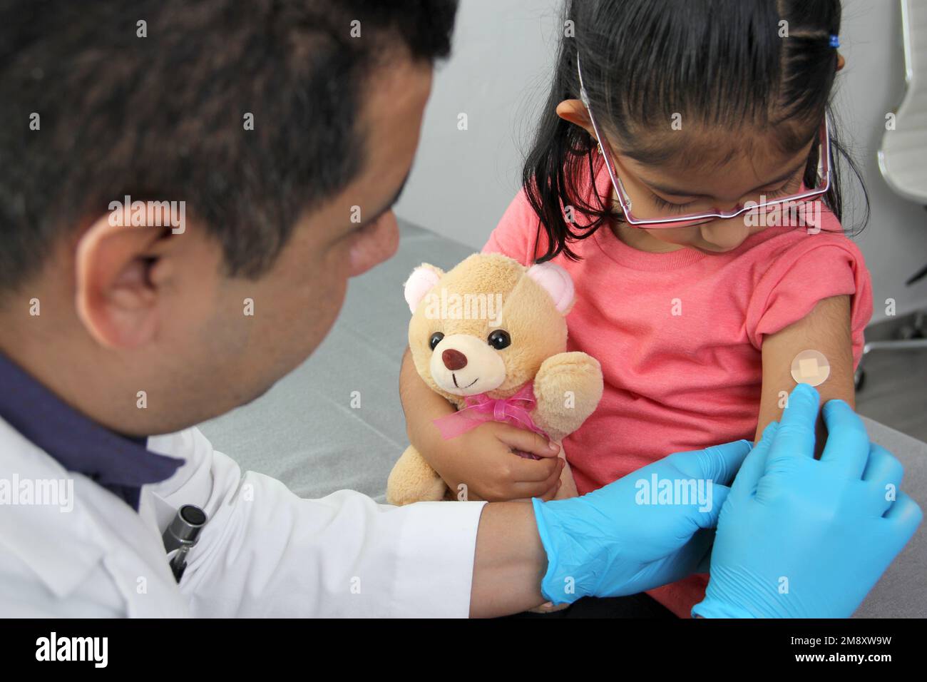 Dark-haired Latino doctor and little girl have a medical consultation in the pediatric office to vaccinate their arm against Covid, chickenpox, diphth Stock Photo