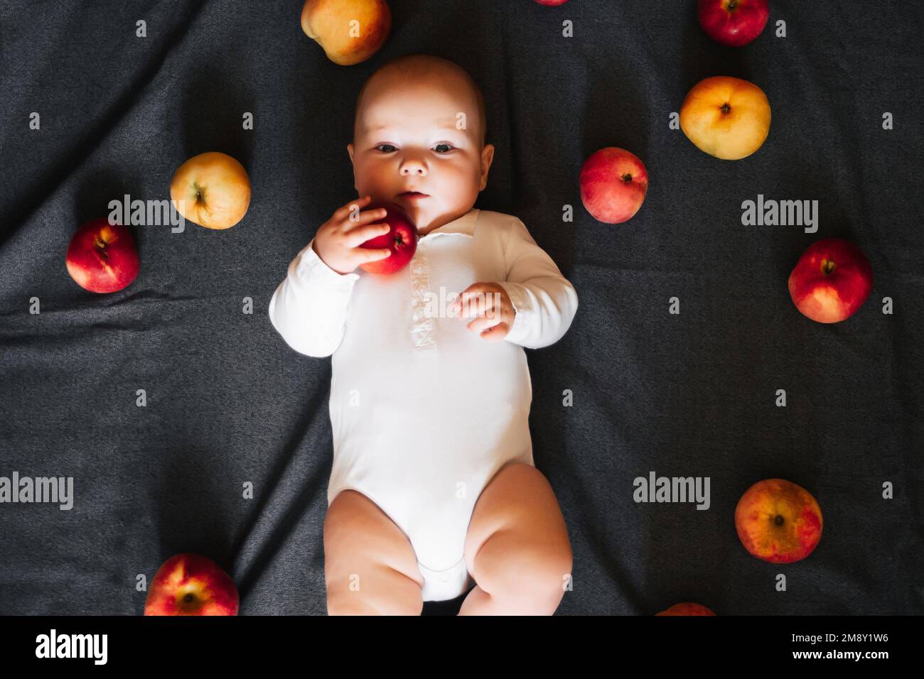 Newborn boy lying with apples. A small child in a white bodysuit on a black background. Top view, flat lay Stock Photo