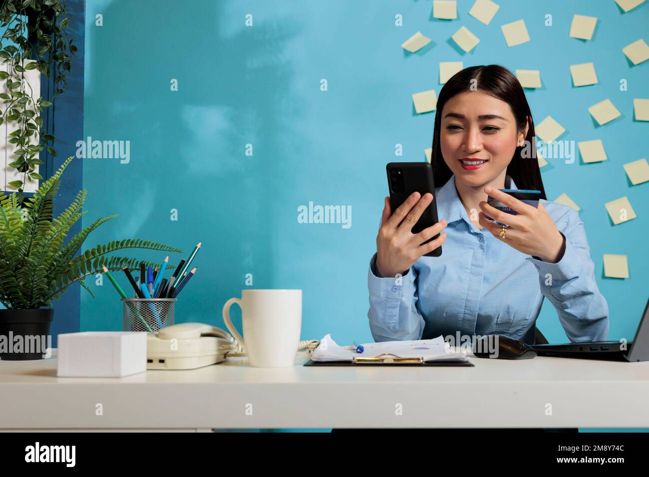 Asian businesswoman holding credit card making financial purchase for business from office. Optimistic female employee making payment transaction at desk using smartphone. Stock Photo