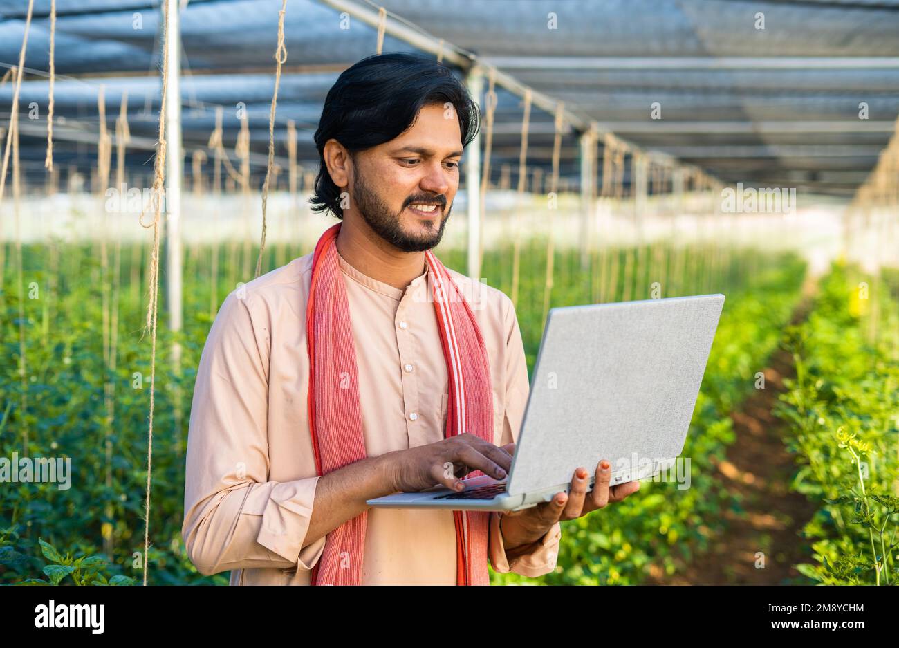 Happy young farmer busy working on laptop at greenhouse - concept of modern farming, technology and development or growth. Stock Photo