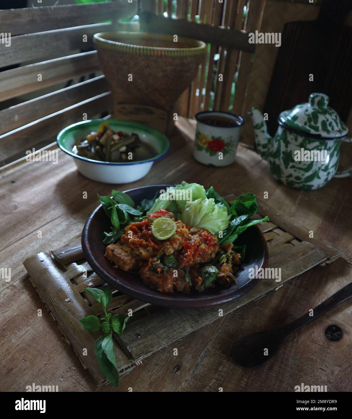 Sambal tempe served with boiled vegetables on a clay plate against a bamboo material background Stock Photo