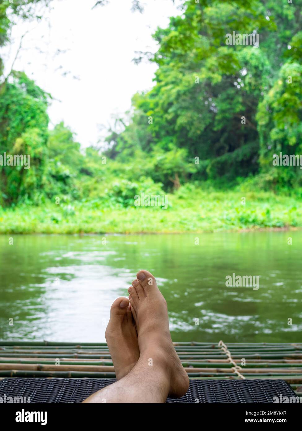 Take a break with relaxing time. The crossed feet of a person resting on a sunbed on a bamboo raft, watching streams in the calm peaceful green forest Stock Photo