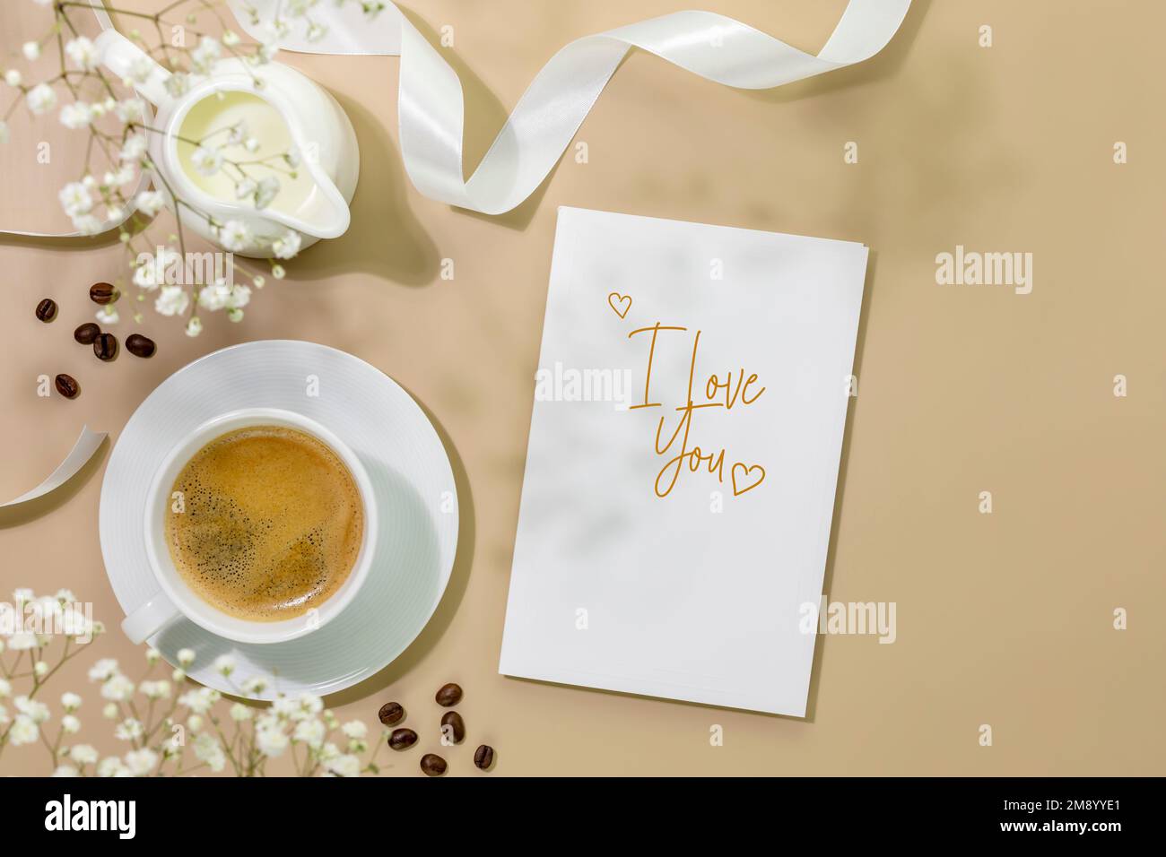 Valentines day flat lay with cup of natural coffee, creamer with milk, coffee beans and gift card with text, Ilove you on beige background with white Stock Photo