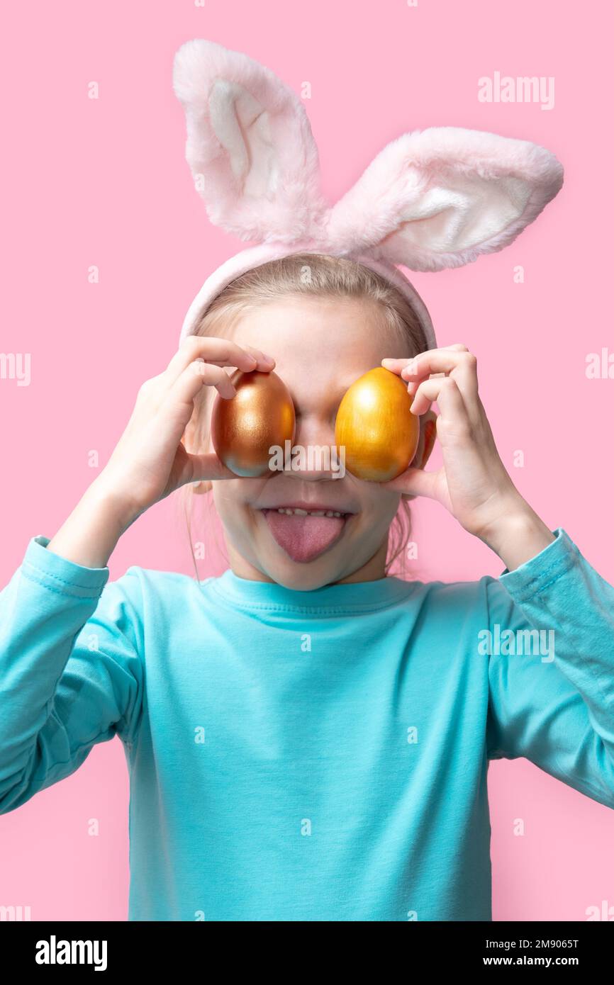A cheerful happy girl in a blue sweater and with bunny ears holding colorful Easter eggs on a pink background, vertical frame. surprised bunny kid. ha Stock Photo