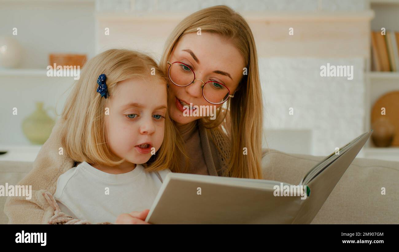 Caucasian mother in eyeglasses with little small cute daughter preschool child girl baby reading book in living room at home mom with kid read Stock Photo