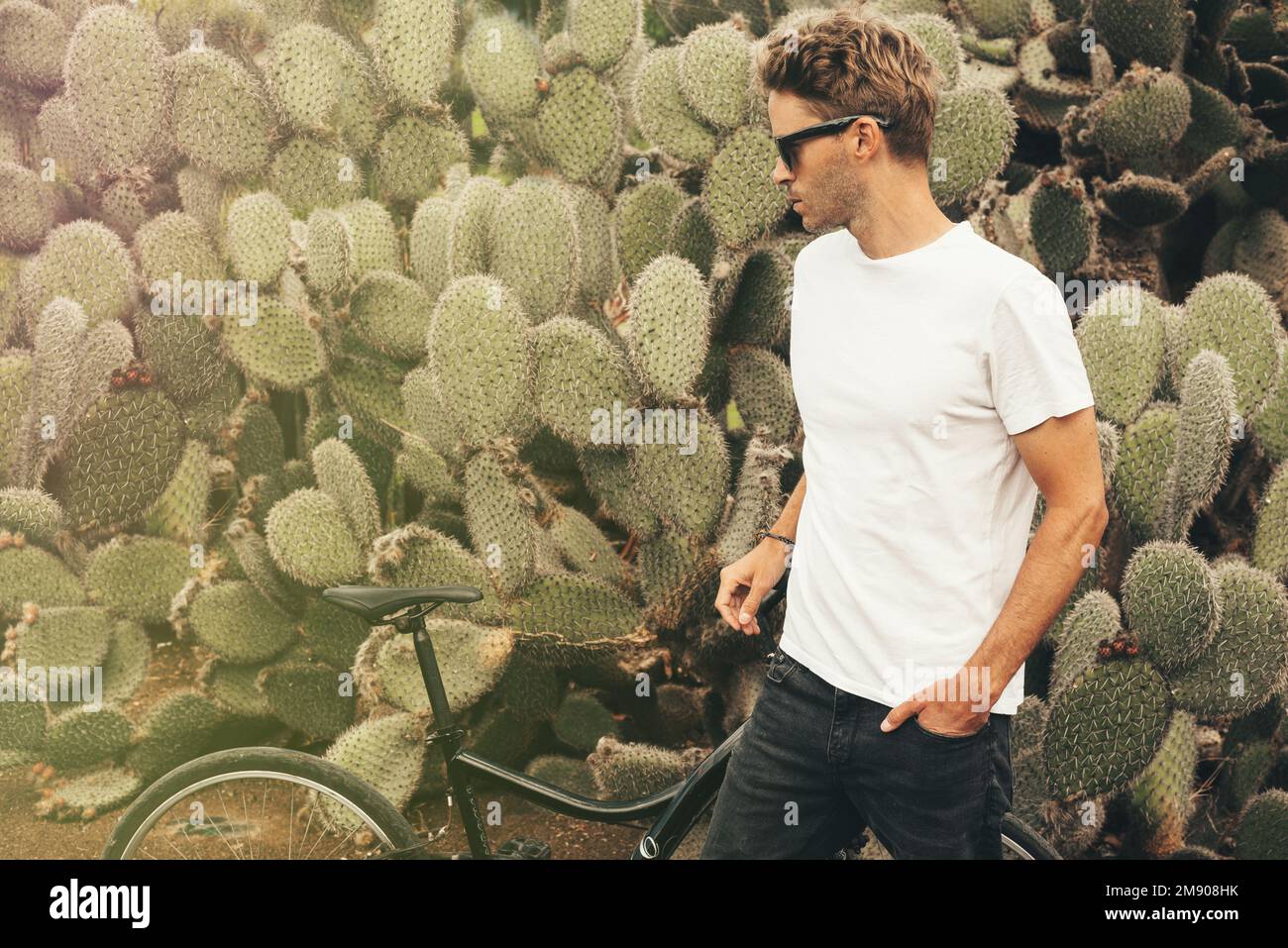 Blond hipster male model wearing sunglasses posing near a big cactus next to his bike. Stock Photo