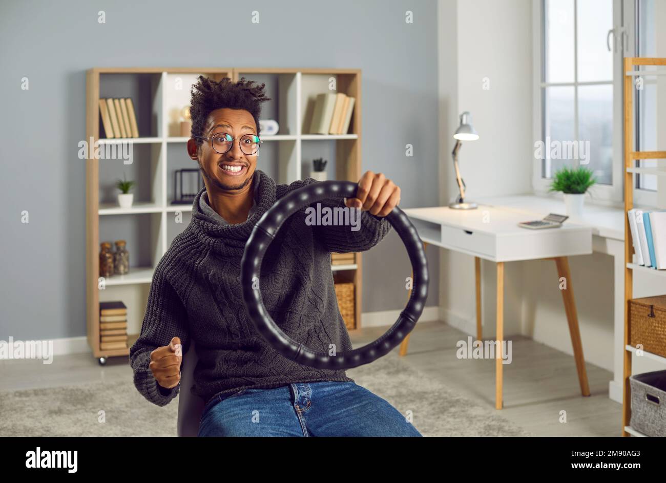Young funny excited indian guy keeping steering wheel sitting on a chair at home. Stock Photo