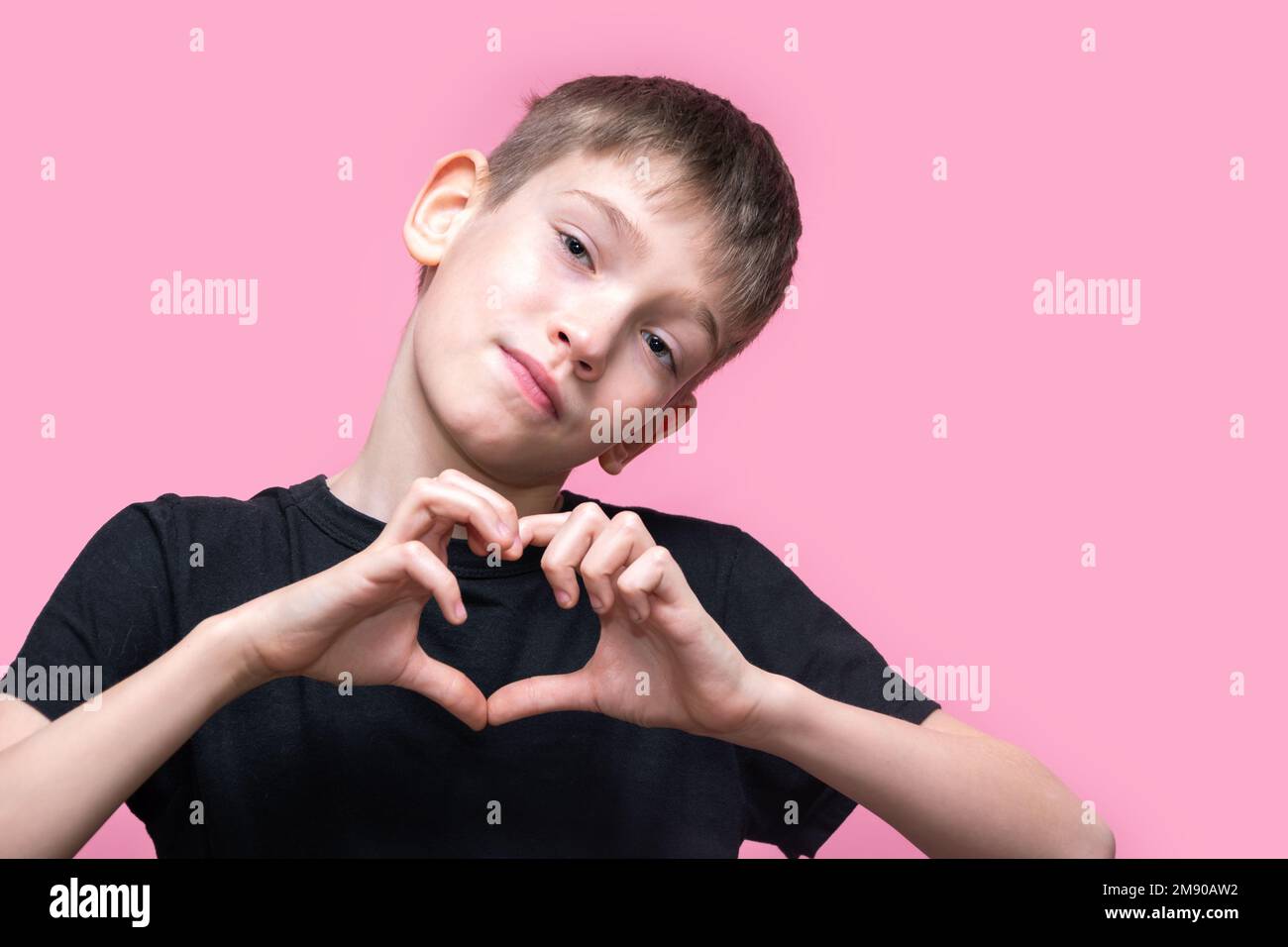 A teenage boy in a black T-shirt makes a heart shape with his hands and smiles on a pink background. Love, relationships. valentine's day. Stock Photo
