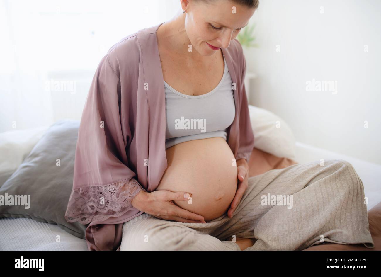 Happy pregnant woman stroking her belly, sitting on bed. Stock Photo