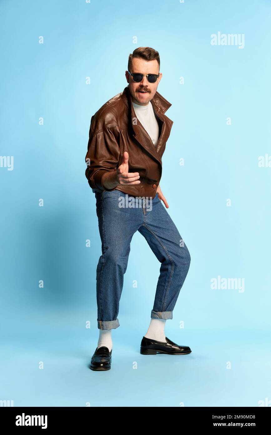 Portrait of brutal, handsome man in jeans, leather jacket and sunglasses posing, dancing over blue studio background. Confidence. Concept of emotions Stock Photo