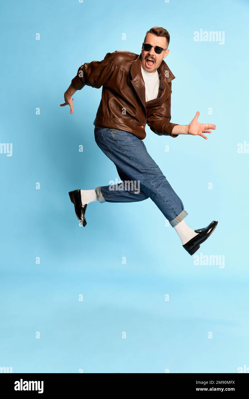 Portrait of brutal, handsome man in jeans, leather jacket and sunglasses posing, jumping over blue studio background. Concept of emotions, lifestyle Stock Photo