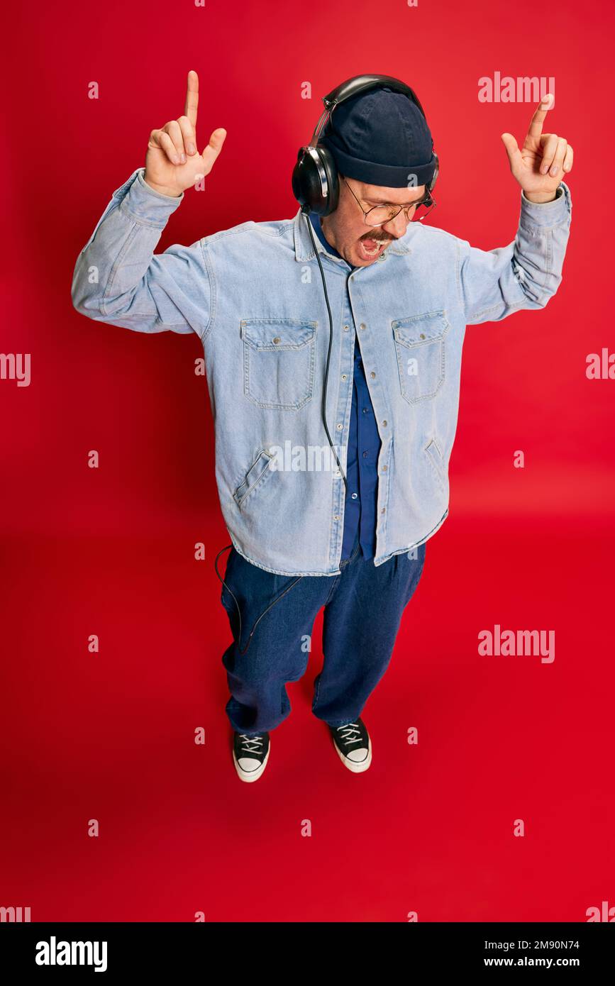 Top view. Portrait of man in stylish modern clothes listening to music in headphones over red background. Concept of modern fashion, lifestyle, music Stock Photo