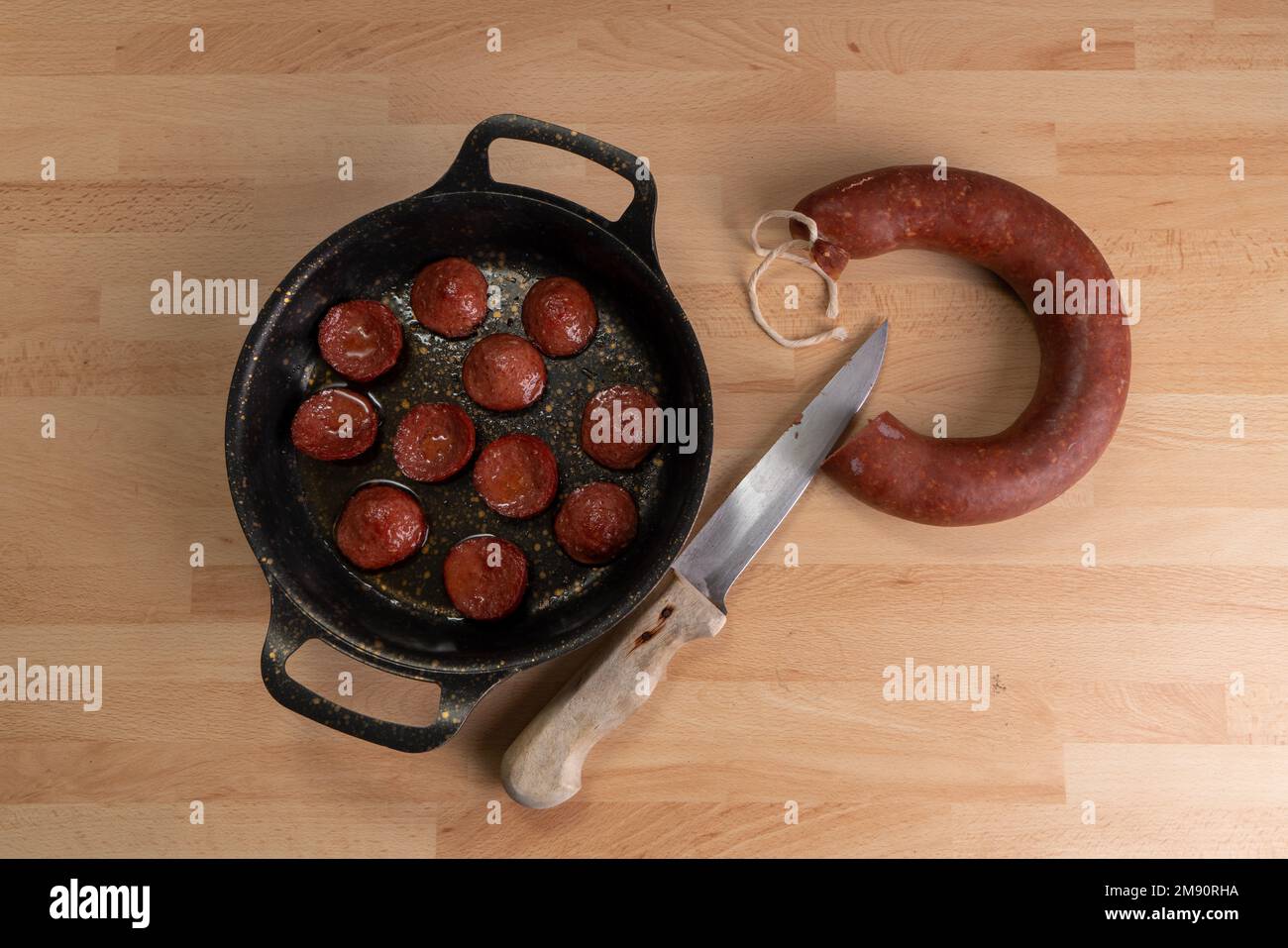 Top view of fried sausage slices in pan. Next to the frying pan are raw sausage and a knife on wooden background. Stock Photo