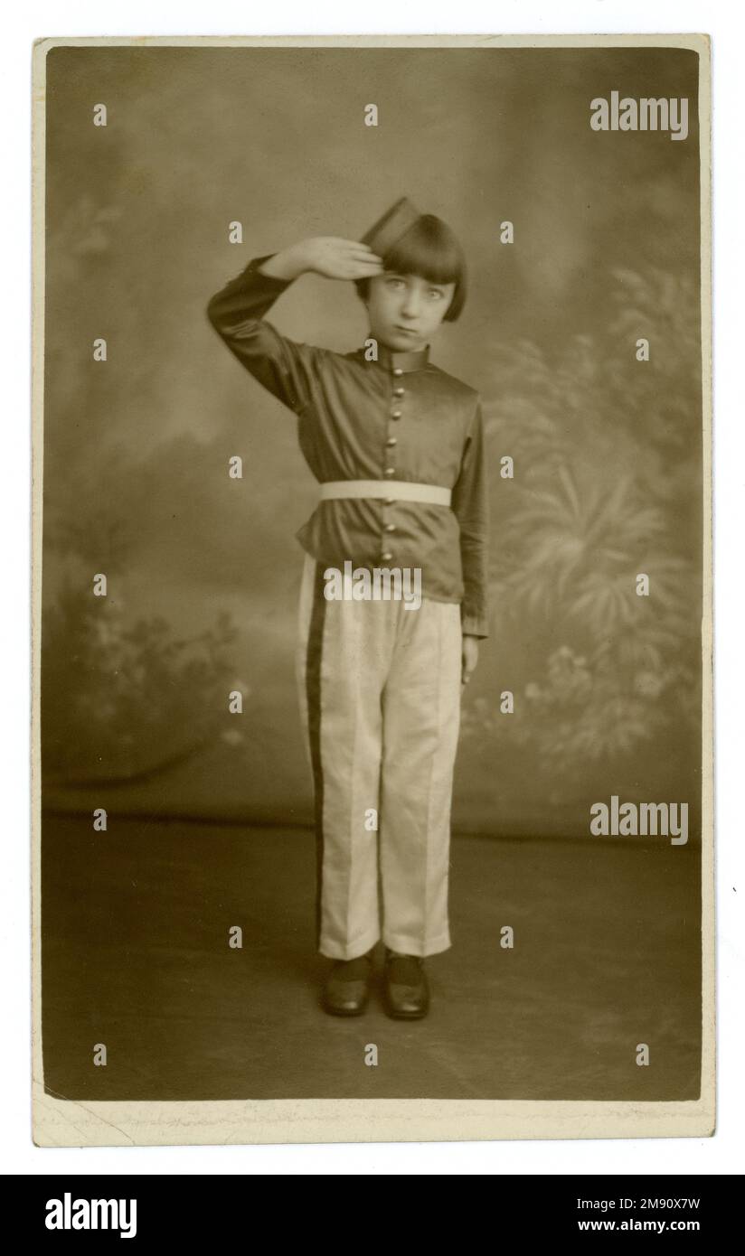 Original 1930's postcard of a cute young girl dressed in the uniform of a youth brass band, military marching band. 1930's childhood. She wears silk trousers and tunic and bellhop hat and is saluting. Postcard is dated 6th Jan 1934 on reverse, studio of Williams Pioneers Studios Ltd. Tooting, Kingsland, Deptford, London. Stock Photo