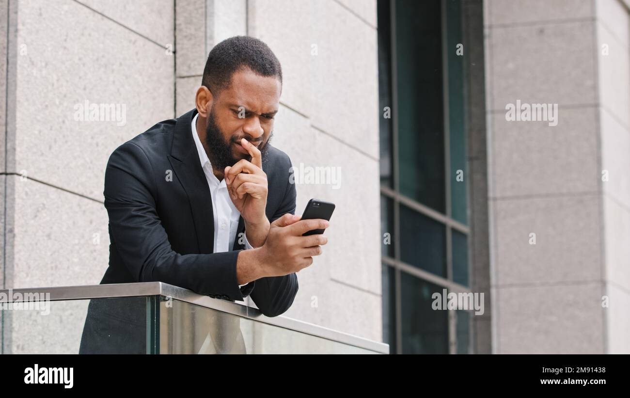 Pensive thoughtful ethnic African American bearded man looking at smartphone screen think about business idea pondering browsing cellphone thinking Stock Photo