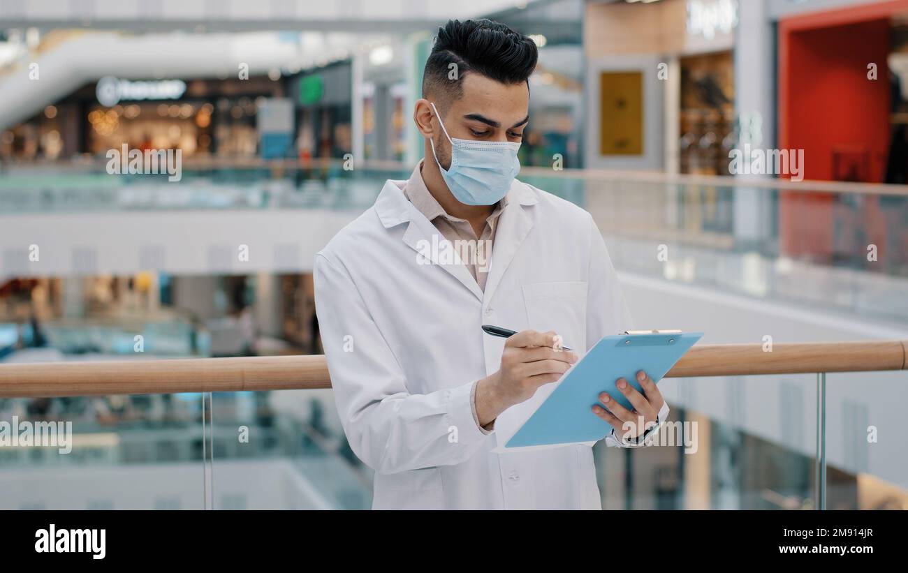 Ethnic Indian medical inspector scientist doctor male worker man writing in papers documents expertise check health safety rules supervisor with Stock Photo