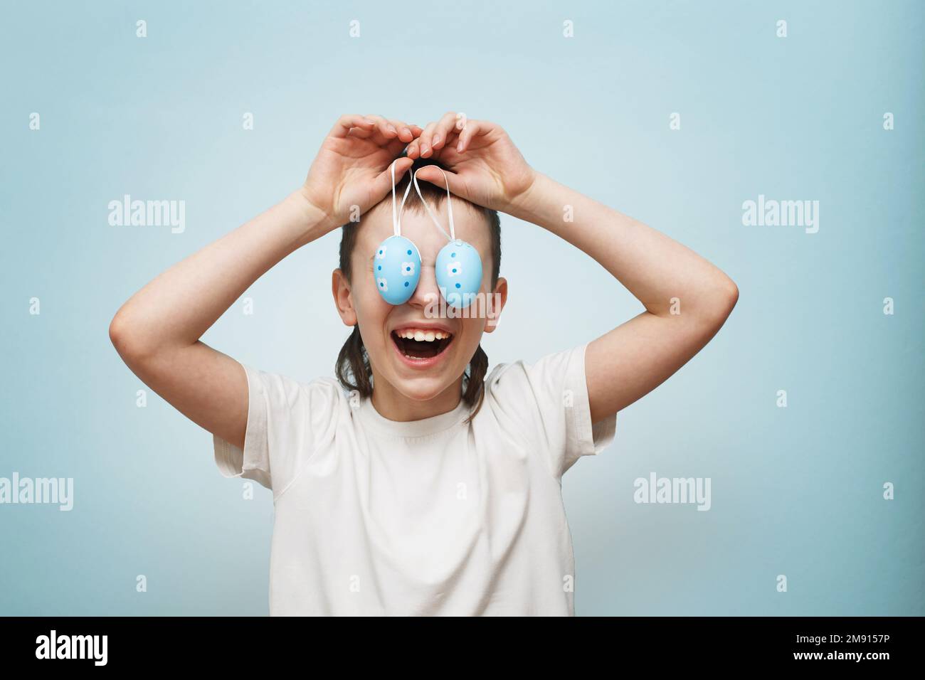young smiling boy holding Easter eggs in his hand and covering his eyes with it on a blue background. Stock Photo
