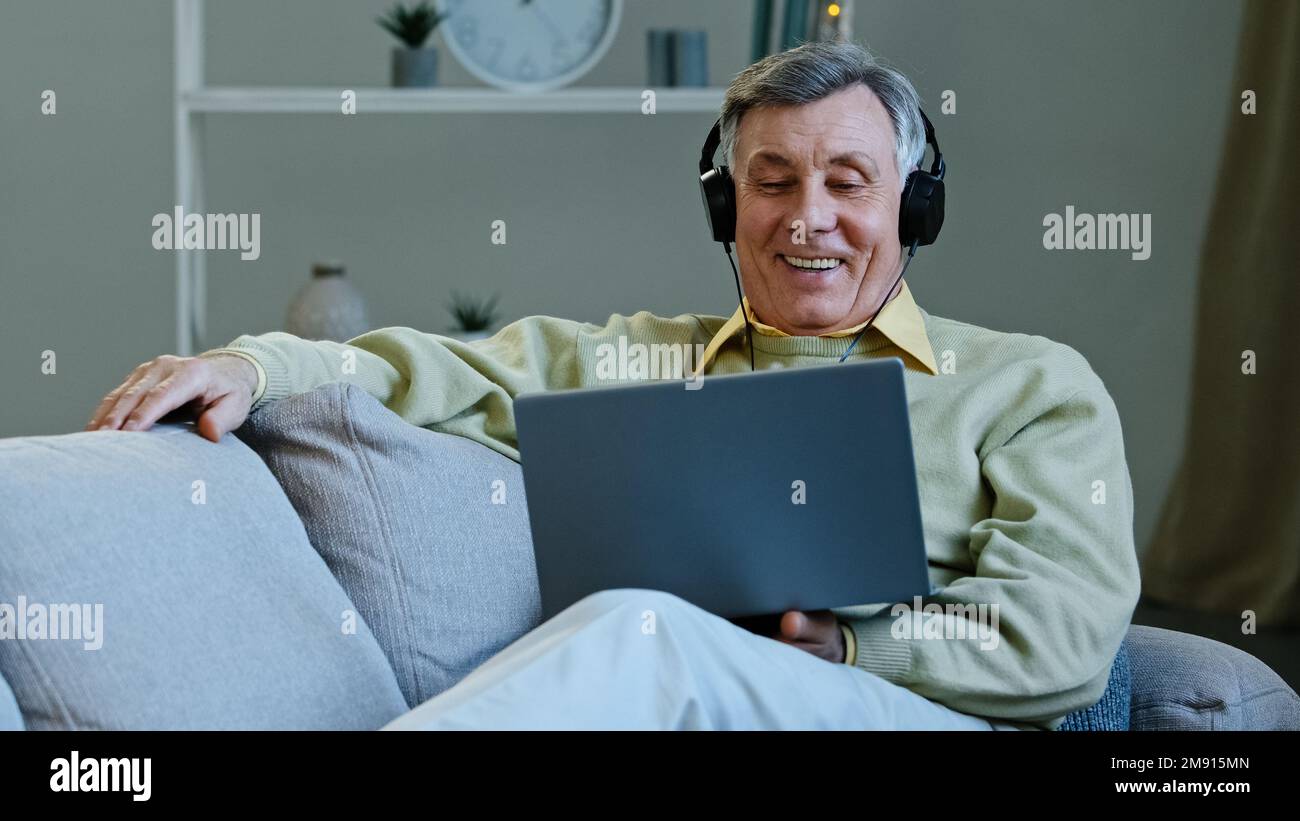 Excited happy aged man watching movie using laptop and headphones sitting lying on comfy couch smiling elderly grandfather relaxing watching comedy Stock Photo
