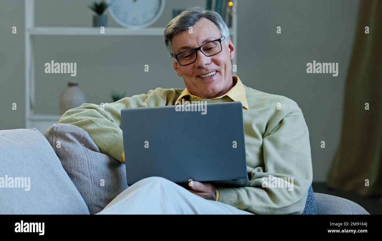 Happy grandfather sit on sofa living room mature elderly caucasian man looking at screen laptop work remotely browses news on internet make online Stock Photo